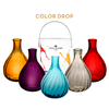 5 small glass bud vases in different colors (orange, purple, blue, gray, and red) in front of a color-splashed paint can that says the brand name "Vista Alegre - 1924" on it -- the image reads "color drop" to show that the color drop bud vases come packaged in a paint can